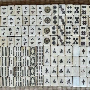 May include: A collection of vintage ivory-colored Mahjong tiles arranged in a grid. The tiles feature Chinese characters, dots, and geometric designs in black ink. The tiles are displayed on a woven surface.