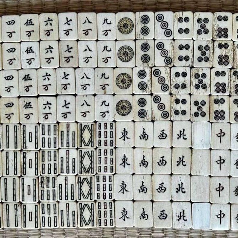 May include: A collection of vintage ivory-colored Mahjong tiles arranged in a grid. The tiles feature Chinese characters, dots, and geometric designs in black ink. The tiles are displayed on a woven surface.
