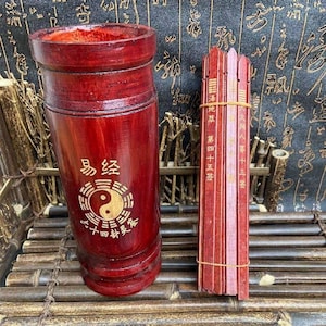 May include: A red wooden divination set featuring a tall, cylindrical container with the Yin and Yang symbol and Chinese characters. Beside it are red divination sticks bound with a rubber band. The set is on a bamboo surface.