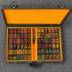 May include: A complete Mahjong set in a decorative case. The wooden tiles feature various symbols and characters in red, green, and black. The case is black with a gold floral pattern and an orange interior.