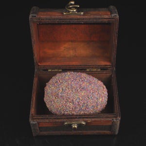 Può includere: Un piccolo forziere di legno aperto con finitura marrone scuro. All'interno, è visibile una grande sfera strutturata e multicolore. La sfera ha un aspetto glitterato con sfumature di viola, blu e oro.