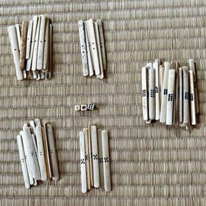 May include: An overhead shot shows a traditional game set, including ivory-colored sticks and three dice. The sticks are arranged in several groups, some with black dot markings. The game pieces rest on a woven, light brown mat.