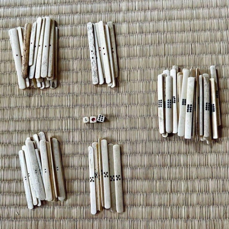May include: An overhead shot shows a traditional game set, including ivory-colored sticks and three dice. The sticks are arranged in several groups, some with black dot markings. The game pieces rest on a woven, light brown mat.