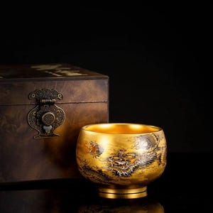 Puede incluir: Un cuenco dorado con un diseño detallado de dragón. El cuenco tiene forma redondeada con una base pequeña. El interior del cuenco también es dorado. En el fondo hay una caja de madera con herrajes metálicos ornamentados.