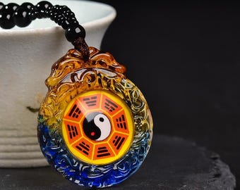 Puede incluir: Un collar con colgante con un colgante circular de vidrio multicolor. El colgante presenta un símbolo de Yin y Yang en el centro y es amarillo, naranja, azul y transparente. El collar tiene una cadena de cuentas negras.