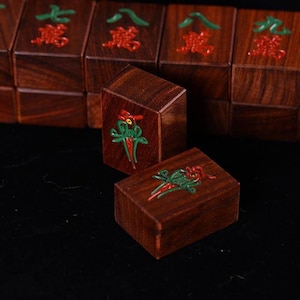 May include: Close-up of wooden Mahjong tiles. The tiles are a rich brown color with red and green embossed designs. The tiles have Chinese characters in red and green. The image shows a few tiles in focus, with others blurred in the background.