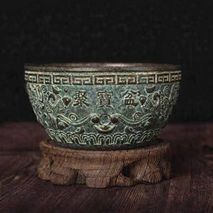 May include: A detailed, antique-style bronze bowl with intricate dragon and geometric patterns. The bowl features Chinese characters and sits on a rustic wooden stand. The bowl has a weathered, green patina.