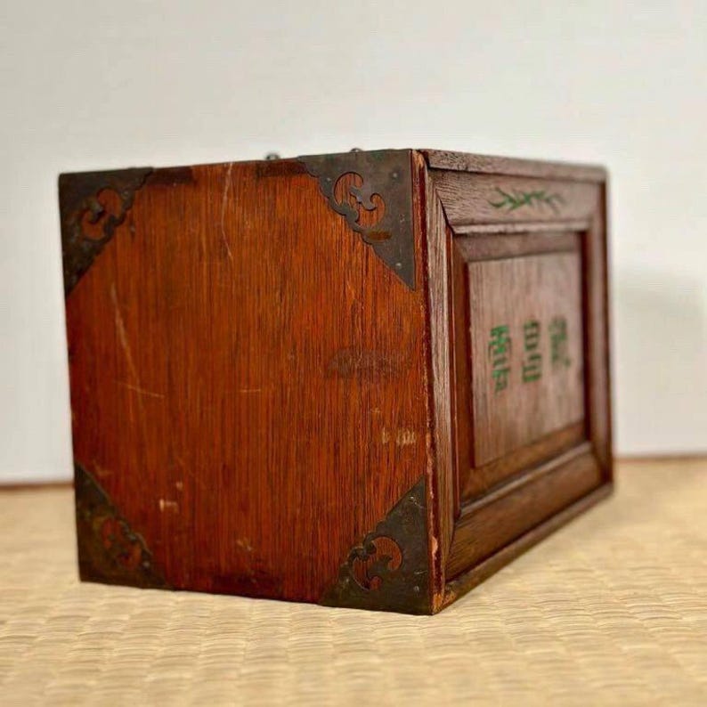 May include: A wooden box with a reddish-brown finish, featuring decorative metal corner brackets. The box has a rectangular shape and appears to be of Asian origin. Green text is visible on the side of the box.