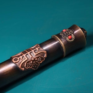 May include: Close-up of a bronze-colored decorative object with intricate floral and geometric patterns. The object has a dark brown cylindrical body with a copper-colored band and accents. Red beads are visible on the end.