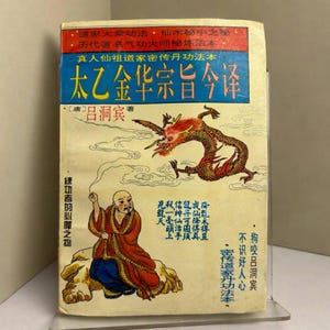 Op de afbeelding: Vintage boekomslag met Chinese tekst en illustraties. Bevat een rode en gouden draak, een geklede figuur en diverse Chinese karakters. De titel staat in grote blauwe letters.