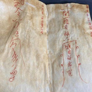 May include: Two-page document with faded, aged paper. Red ink calligraphy and markings cover the pages. The paper has a light brown hue with visible creases. The text appears to be in an Asian language.