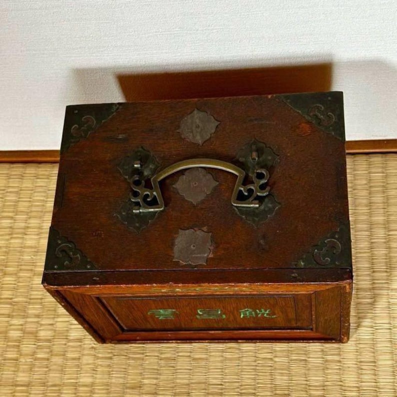 May include: A rectangular wooden box with a dark brown finish, metal corner accents, and a brass handle. The box features decorative metal embellishments and a small inscription on the front. The box is likely used for storage.
