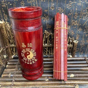 Puede incluir: Un juego de adivinación de madera teñida de rojo. El juego incluye un recipiente cilíndrico con caracteres chinos y un símbolo yin-yang, junto con un paquete de palos de madera. Los palos están atados con una goma elástica y tienen texto chino.
