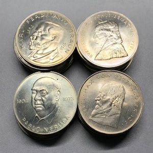 May include: Four stacks of silver coins, each displaying a portrait of a historical figure. The coins feature Karl Ferdinand Braun, Friedrich Schleiermacher, Pablo Neruda, and Max von Pettenkofer. Dates and names are inscribed on the coins.