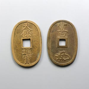 Puede incluir: Dos antiguas monedas japonesas ovaladas con agujeros cuadrados. Las monedas son de color bronce y presentan caracteres japoneses. Una moneda tiene los caracteres "天保通寶" y la otra "當百".