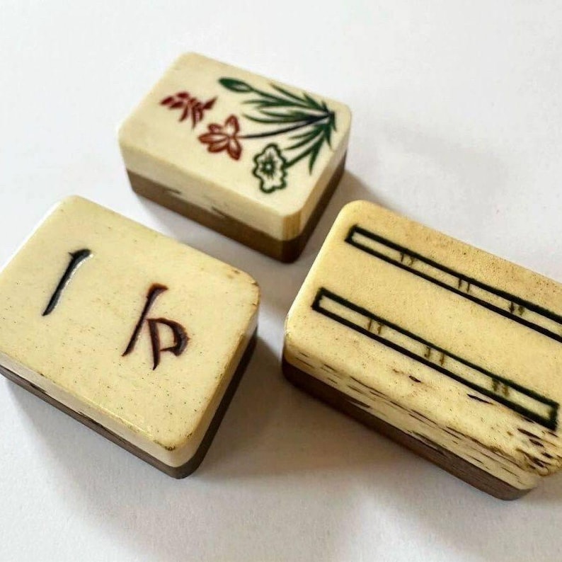May include: Three vintage Mahjong tiles. The tiles have a cream-colored top with brown wooden bases. One tile has black characters, another has a floral design in red, green, and cream, and the third has black lines.