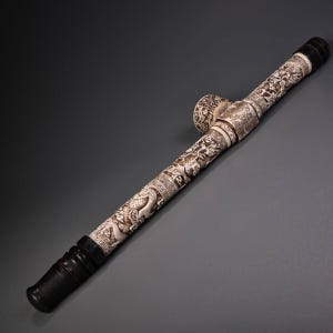 May include: A long, cylindrical smoking pipe featuring intricate, carved off-white relief patterns of dragons and geometric borders. The pipe has dark, smooth, polished black ends and a circular bowl section positioned along the main shaft.