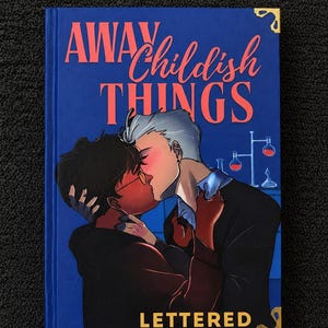 Pu&ograve; includere: Un libro con copertina rigida blu intitolato AWAY Childish THINGS LETTERED con l'illustrazione di due persone che si baciano. Una ha capelli scuri e occhiali, l'altra capelli chiari. Uno sfondo di laboratorio con becher e angoli dorati.