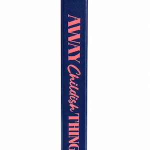 Pu&ograve; includere: Un dorso di libro blu scuro con la parola LETTERED stampata in piccole lettere maiuscole rosa nella parte superiore. Sotto, il titolo AWAY Childish THINGS &egrave; stampato in grandi lettere rosa. Un piccolo segnalibro a nastro rosso sporge.