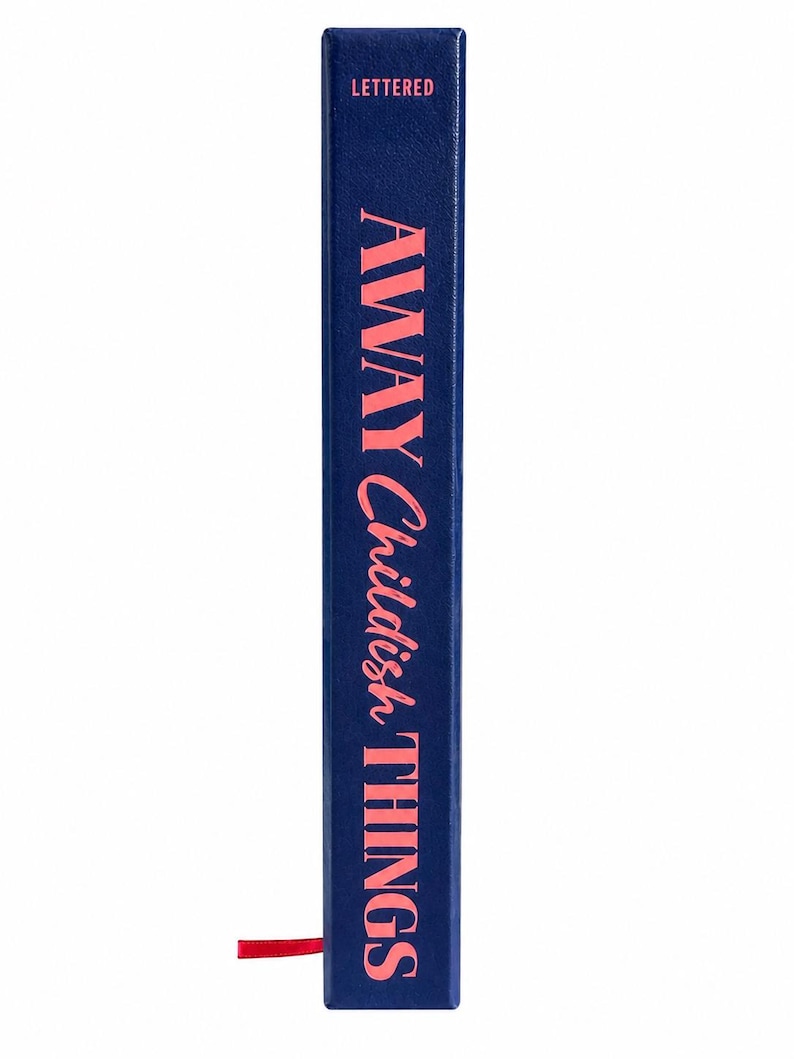 Pu&ograve; includere: Un dorso di libro blu scuro con la parola LETTERED stampata in piccole lettere maiuscole rosa nella parte superiore. Sotto, il titolo AWAY Childish THINGS &egrave; stampato in grandi lettere rosa. Un piccolo segnalibro a nastro rosso sporge.