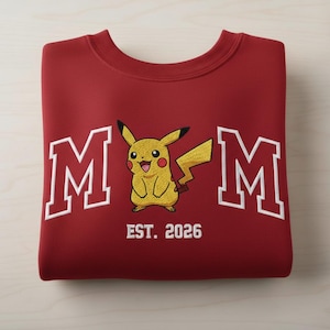 May include: A maroon sweatshirt with white embroidered text and a yellow Pikachu design. The word "MOM" is on either side of the Pikachu, with "EST. 2026" below the character.