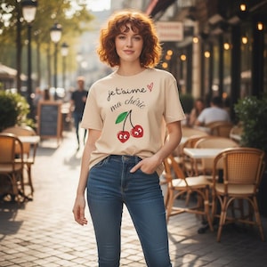 Puede incluir: Camiseta beige con la frase francesa "Je t'aime ma chérie" y un gráfico de cerezas. La camiseta se combina con vaqueros azules. La imagen está tomada al aire libre, con un fondo borroso de una calle con cafés.