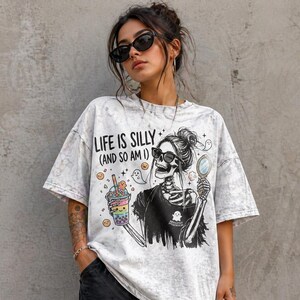 May include: A light gray, oversized t-shirt with a graphic of a skeleton wearing sunglasses, holding a mirror and a colorful drink. The text "LIFE IS SILLY (AND SO AM I)" is printed on the shirt.
