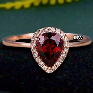 May include: A rose gold ring with a pear-shaped, deep red gemstone and a halo of small, clear stones. The ring has a slender, polished band. The initials "JBW" are visible on the band. The ring is set against a dark background.