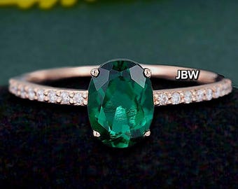 May include: Rose gold ring featuring a large, oval-cut emerald gemstone. The band is adorned with small, clear gemstones. The initials "JBW" are visible on the band. The ring is set against a dark background with green and yellow foliage.
