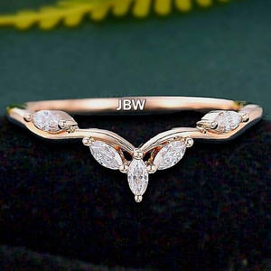 Puede incluir: Anillo de oro rosa con un diseño de racimo de diamantes talla marquesa. El anillo presenta una banda delicada y una forma curva, con las iniciales "JBW" visibles en la banda. El anillo está sobre un fondo oscuro.