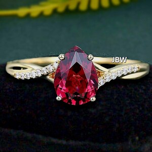 May include: A gold ring with a pear-shaped, deep red gemstone as the focal point. The band features a twisted design with small, clear accent stones. The ring is set against a dark background, highlighting its sparkle.
