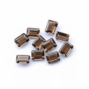 May include: A scattering of rectangular, faceted smoky quartz gemstones. The gems are a warm, translucent brown colour and are on a white background. The cut of the stones is an emerald cut, showcasing their clarity and colour.