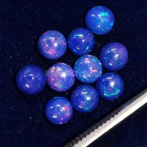 May include: A collection of ten round, polished blue opal gemstones. Each stone has a vibrant, iridescent play of color, with flashes of pink and green. The opals are displayed on a dark blue surface.