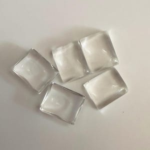 May include: Five small, clear, rectangular glass cabochons with smooth, rounded edges and a flat surface. These transparent pieces are arranged in a scattered formation, reflecting light across their polished, glossy surfaces and clear glass bodies.