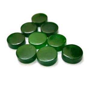 May include: A collection of ten polished, round, dark green jade stones. Each stone is a flat disc shape, with a smooth, reflective surface. The stones are scattered on a white background, showcasing their natural color and texture.