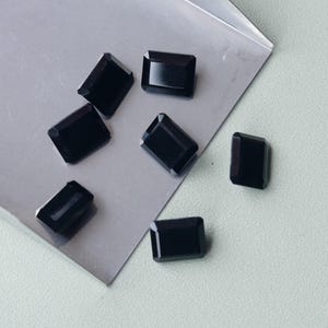 Puede incluir: Siete gemas negras rectangulares con un acabado pulido están dispuestas sobre una superficie gris claro. Las gemas tienen un corte esmeralda clásico, mostrando su color oscuro y opaco. La disposición sugiere una colección de piedras sueltas.