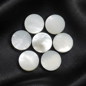 May include: Seven round, white mother-of-pearl discs arranged in a circular pattern on a black background. The discs have a pearlescent sheen, reflecting light. The overall effect is elegant and natural.