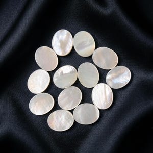 May include: A collection of twelve oval-shaped mother of pearl cabochons. Each piece features a smooth, polished surface with a natural iridescent white finish, displaying subtle variations in luster and light reflection across the curved forms.