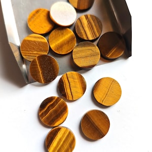 May include: A selection of circular tiger's eye gemstones in gold and brown hues. The gemstones are presented in a metal tray against a white backdrop. The stones have a polished, smooth surface.