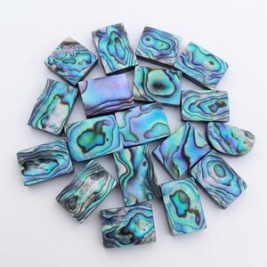 Könnte beinhalten: Eine Sammlung rechteckiger Abalone-Muschel-Cabochons mit irisierenden Mustern in Blau-, Grün- und Violetttönen. Jedes Stück weist eine einzigartige, wirbelnde organische Textur mit einer polierten, reflektierenden Oberfläche auf.