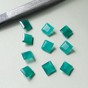 Op de afbeelding: Een verzameling vierkant geslepen, smaragdgroene edelstenen. De edelstenen liggen op een lichtgekleurd oppervlak, met een pincet in de linkerbovenhoek. De edelstenen hebben een gepolijst, reflecterend oppervlak.
