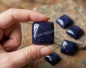 May include: A square blue sandstone cabochon with a glittery, star-like appearance and a smooth, polished surface. The stone features the text WILDGEMCREATIONS printed across its center. Several additional matching blue sandstone pieces are visible.