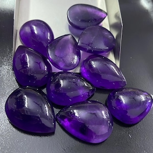 May include: A collection of polished, teardrop-shaped amethyst gemstones. The gems are a deep, rich purple color and have a smooth, reflective surface. They are arranged in a small, silver-colored tray, showcasing their natural beauty.