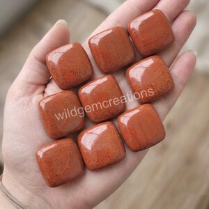 May include: Eight polished, square-shaped red jasper stones with smooth, rounded edges and a glossy finish. The stones feature a deep terracotta color with subtle dark speckling throughout. The text "wildgemcreations" is overlaid across the center.