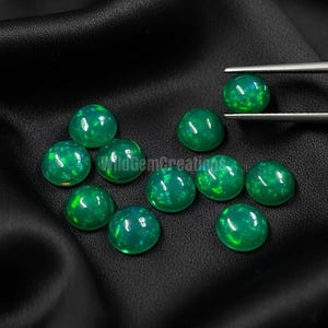 May include: A collection of round, polished green gemstones with iridescent flashes, displayed on a dark, textured surface. The gemstones are of varying sizes and are arranged in a scattered pattern. A pair of tweezers holds one of the gemstones.