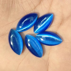 Op de afbeelding: Zes ovale, gepolijste edelstenen in verschillende blauwtinten zijn in een cirkelvormig patroon gerangschikt. Elke steen heeft een prominent, lichtgekleurd kattenoogeffect dat door het midden loopt, wat een opvallend visueel contrast creëert. De edelstenen worden tegen een neutrale achtergrond gepresenteerd.