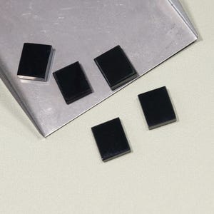 May include: Five small, rectangular, black gemstones are arranged on a silver tray. The gemstones have a polished, reflective surface. The tray sits on a light-coloured surface.