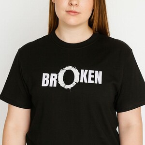 May include: Black crew neck t-shirt with the word "BROKEN" printed in white. The "O" in "BROKEN" has a distressed, cracked design. The model is wearing the shirt.