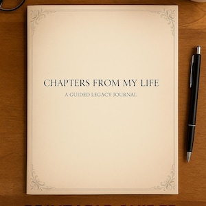 May include: A cream-colored journal with the text "CHAPTERS FROM MY LIFE" and "A GUIDED LEGACY JOURNAL". The journal has decorative corner designs and is on a wooden surface. A black pen and glasses are also present.