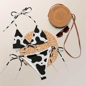May include: A black and white cow print bikini set with tie closures. The bikini is displayed with a woven straw mat, sunglasses, and a round woven purse with a brown strap. The bikini top and bottom are patterned with black spots.